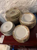 Stacked multiple sets of china plates and bowls on a wooden surface against a wall, showing three distinct groups with different floral and gold rim designs.