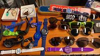 Wide view showing multiple wristwatches in various colors and styles, alongside several pairs of sunglasses spread on a wooden surface.
