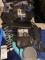 Lot displayed with all items on a blue surface including Olympus OM-1 with Sigma lens, GoPro Hero in waterproof case, Nikon CoolPix 7600 camera and Joby Spider tripod