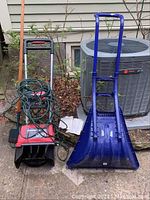 Photo showing Toro Power Curve electric snow shovel in red and black, blue plastic shovel, and red hand truck against house wall with air conditioning unit.