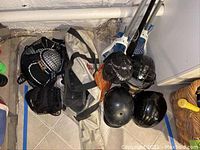 Photo showing multiple black helmets, black protective pads, a white and black sports bag and several lacrosse sticks with white heads and blue details arranged on floor against wall