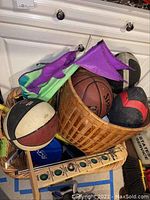 Photo showing woven basket filled with multiple basketballs, footballs, and fabric item