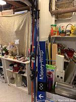 View of skis, ski poles, and blue Misery Whip ski trail sign leaning against basement wall with other household items.
