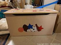 Front view of the veneer toy box showing sports-themed painted decorations including basketball, football, soccer ball, baseball bat and ball, blue and red stars.