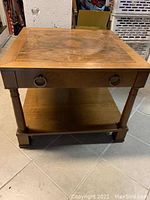 Front and side view of wooden side table with drawer pulls and lower shelf on tiled floor.