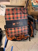 Top view showing Forever Young red and black plaid bag with logos on black band wrapped around, resting on an orange backpack.