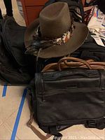 Photo of wool felt cowboy hat with feather band atop black leather wheeling briefcases and backpacks on floor.