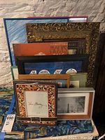 Group of assorted framed pictures in multiple styles and sizes, stacked together showing variety of frames: ornate gold, blue glass, simple green, others.