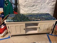 Full front view of the white wooden bench showing its cushioned top with a multicolor abstract pattern, two side doors with black knobs, and open middle shelves.