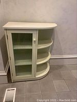 White corner cabinet with glass door and three curved side shelves, placed against wall on tiled floor