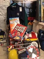 Photo of assorted Wheaties and Flutie Flakes cereal boxes with men's sports jersey hanging behind them and various sports memorabilia.