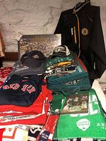 Lot laid out showing multiple folded T-shirts in Red Sox, Bruins, and Celtics colors, Bruins framed Stanley Cup plaque, hats, rally towels, and seat cushions.