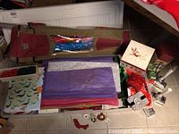 Photo showing multiple rolls of Christmas wrapping paper in a carrying case along with decorative boxes, green and red string, and small ornaments scattered on table