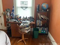 Desk with chair, covered with books and office items, next to a bookshelf and a window.