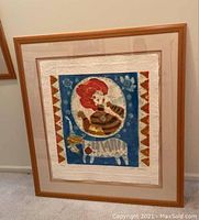 Framed signed and numbered relief print titled 'L'Amazone', edition 18/75, with a stylized figure with red hair and an animal, set in a wooden frame with beige matting.