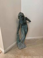 Full view of the metal sculpture of a nude woman holding a cloth, standing on a base against a white wall on carpeted floor.