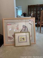 Two framed art pieces leaning against a wall: larger Native American watercolor in light wood frame and smaller print with gold ornate frame.
