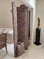Side angle view of the upholstered three-panel room screen with four pillows placed at the base. The fabric is padded and patterned with muted earth tones and abstract leaf/feather designs.