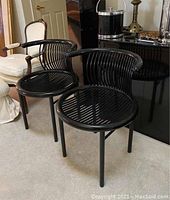 Two black tubular metal accent chairs shown side by side in a living room setting with carpeting and other furniture around.