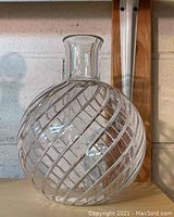 Front view of clear, round crystal vase with spiral etched pattern and short pinched neck on light-colored surface against brick wall background.