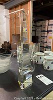 Clear crystal obelisk standing on a table with various cups and glassware around it in a storage or basement area.
