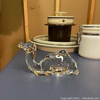Clear Baccarat crystal camel figurine on a blue surface with background household items.
