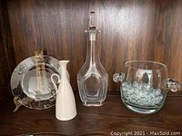Four-piece set on wood shelf showing Lenox white ewer, Tiffany & Co. crystal plate with easel, Orrefors decanter, and glass ice bucket with marbles.