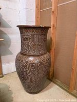 Full view of the tall brown vase with white intricate carved geometric and spiral designs standing upright in a corner.