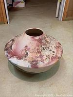 Side view of a rounded ceramic planter or vase with a distressed finish and mottled pink, white, and brown coloring on a concrete floor.