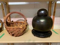 Photo showing the urn on pedestal and the woven basket side by side on shelf, clear view of both items' shape and textures