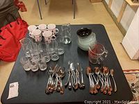 Table displaying all items in the lot: coffee mugs, assorted glasses, flatware, measuring cups, bowls, and pitcher.