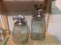 Two large hand blown glass canisters with pewter lids featuring a fish and a chicken on top, showing full view and relative size, with ribbed vertical texture on glass.