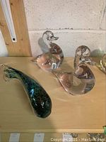 Overall view of two clear glass ducks and one green and blue glass fish paperweight on a wooden shelf with concrete background.