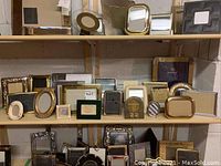 Photo showing three shelves displaying a mixture of picture frames including oval, rectangular, and round shapes in metal, glass, ceramic and fabric. Frames vary in color with gold and silver tones prominent. Some discoloration and chips visible on some frames.