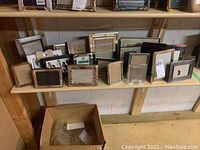 Wide view of multiple picture frames arranged on a wooden shelf in basement. Various sizes and materials including metal, glass, ceramic, fabric present.