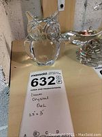 Clear Daum crystal owl figurine sitting on a shelf next to other glass items with tag showing 3.5x3 inch measurement and label indicating Daum Crystal Owl.