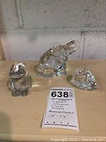Clear crystal rabbit, elephant, and hedgehog figurines on wooden shelf with handwritten lot tag.