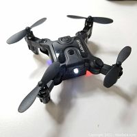 Photo of a powered-on LOLY V2 Mini drone showing black folding arms and propellers, central power button, and LED lights active.