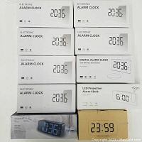 Stack of 10 alarm clocks in their white and beige boxes showing product images and digital times 20:36 and 23:59