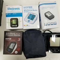 Photo showing 6 blood pressure monitors. Four boxed new devices from brands HOMIEE, EHPOW, and another electronic arm monitor, plus two monitors out of their boxes with cuffs attached.