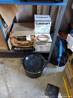 Overview of kitchenware set including the Cuisinart boxed appliance, black stock pot with lid, and roasting pan on floor beneath shelving unit.
