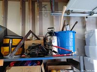 Wide view of shelf showing various tools including metal angle ruler, sprayer, blue bucket, and axe handles