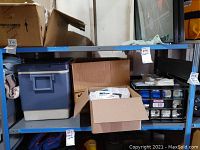 Shelf with a large blue cooler, cardboard boxes, and a plastic rack containing small containers and packs.