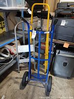 Blue metal hand truck upright with strap and black handles, showing floor and surrounding shelving