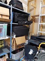 Black rolling suitcases and duffel bags stacked on a gray metal shelf along with a cardboard box and plastic bins underneath.