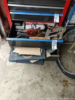 Overall view of open toolbox drawers containing various hand tools including wrenches, hand crank drill, planer, and sanding materials. Shelving and floor visible.