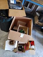 Open box with visible stainless steel boiler pan on top, various packed kitchenware, and colorful plastic plates and cups to the side.