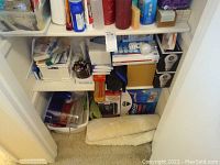 Wide view of closet shelves with household supplies including boxes of light bulbs, bottles, and packing materials on bottom shelf.