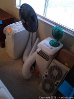 Two white portable air conditioners side by side with black pedestal fan behind and green desk fan on one unit, white box fan in front