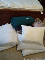 White comforters, green wool blanket, multicolored afghan, and three pillows arranged on carpeted floor next to bed